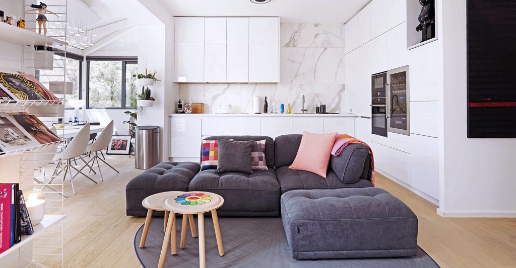 Vue de face du canapé gris, décoré avec plusieurs coussins et une couverture pliée, de la pièce à vivre de Jo Yana, avec la cuisine blanche en L et la table à manger en arrière plan.