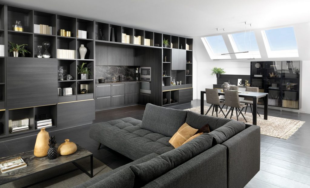 Vue de biais de la cuisine ouverte Arcos, coloris noir effet bois, avec une fenêtre sur la sous pente, une table et des chaises dans le fond et le salon en premier plan.