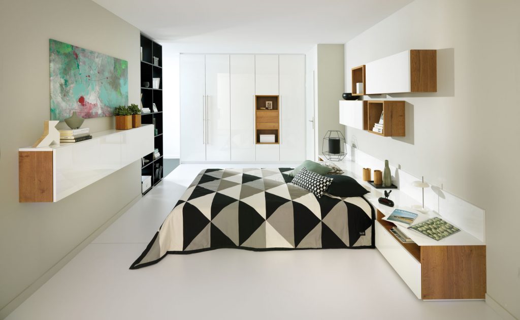 Vue d'ensemble d’une chambre de style contemporain, avec le lit et ses meubles au premier plan, le placard dressing en arrière plan, un meuble suspendu sur la gauche, et des caissons sur la droite, tous dans les coloris blanc et décor bois.