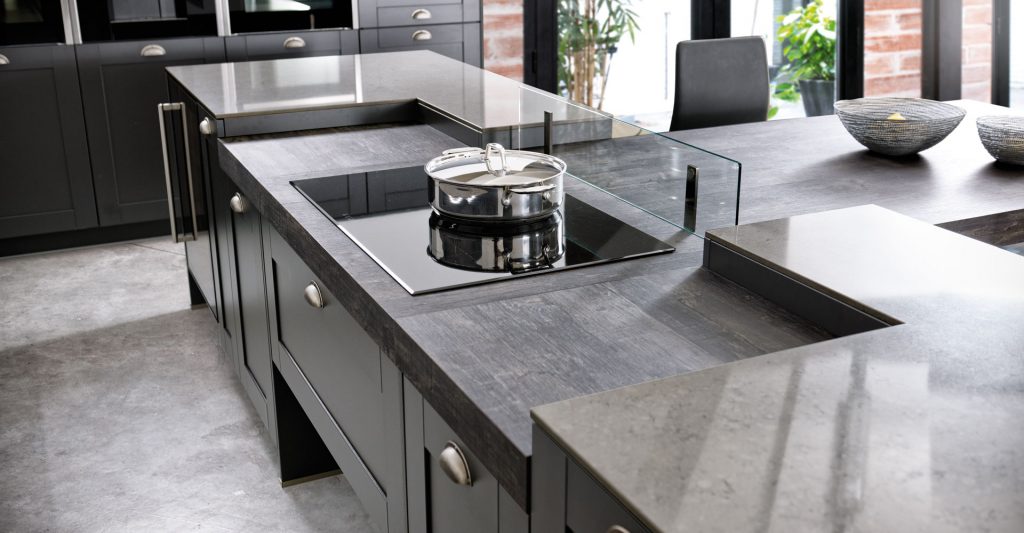Vue par au-dessus de la zone cuisson du plan de travail gris avec la plaque de cuisson et le verre anti projection dans une cuisine ouverte Schmidt, gamme Frame.