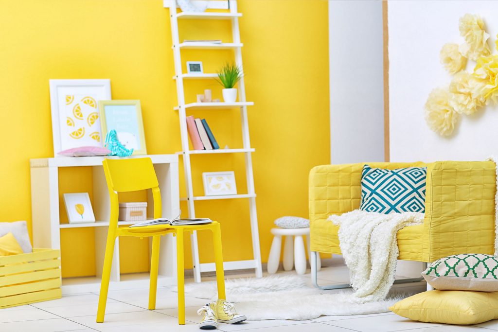 Chambre avec un mur jaune, deux étagères blanches, une chaise et un fauteuil jaunes et des éléments de décoration en bleu, jaune et blanc.