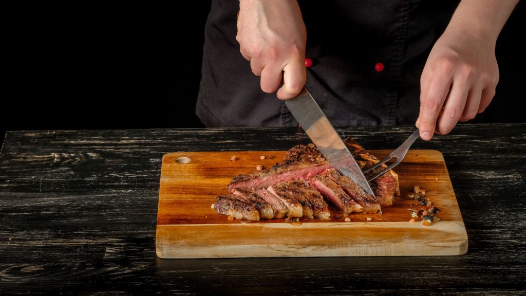 Gros plan sur une planche en bois sur laquelle on voit les mains d’un chef en train de couper un steak cuit.