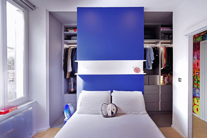 Vue du dressing de Jo Yana en arrière plan, caché en partie par le lit avec un mur bleu comme tête de lit au milieu.